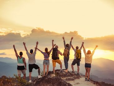 Group of happy friends stands with raised arms at sunset mountain and enjoys sunset