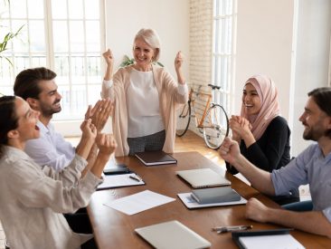 Overjoyed middle aged female team leader celebrating corporate success with young multiracial colleagues at business meeting in office. Happy mixed race teammates succeed in profitable project.